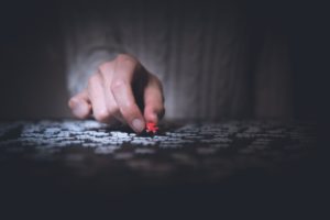 Someone places a red coloured puzzle piece onto a table of all white puzzle pieces